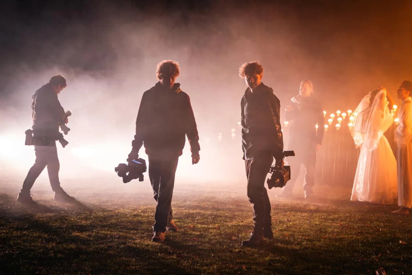 Crew met camera's op een nachtelijke set, tegenlicht door rook en fakkels, figuranten in het wit op de achtergrond
